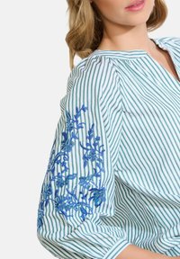 Blusa a righe bianca e verde con ricami floreali blu sulle maniche. Presenta un colletto a V e dettagli arricciati lungo il decolleté.