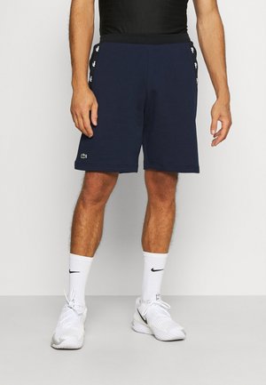 Navy blue athletic shorts with a relaxed fit, elastic waistband, and white brand accents on the side. Paired with white sneakers and socks.