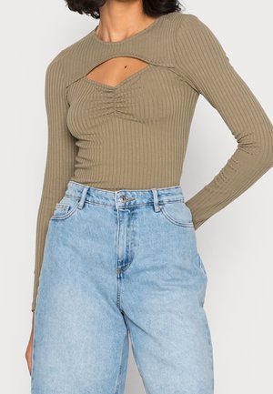 Femme portant un haut côtelé à manches longues couleur olive avec une découpe à l'avant et un jean taille haute bleu clair.