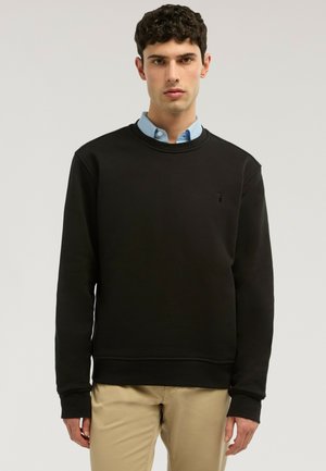Jeune homme portant un sweat-shirt noir à col rond par-dessus une chemise bleue à col, avec un pantalon beige, debout devant un fond clair uni.
