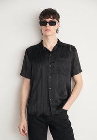 Camicetta nera a maniche corte in satin con righe verticali, chiusura con bottoni sul davanti e una tasca sul petto. Indossata con occhiali da sole neri rettangolari.