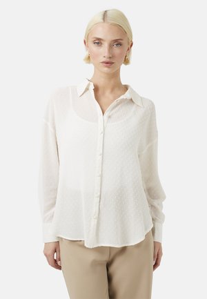 Jeune femme aux cheveux blonds courts portant un chemisier blanc texturé boutonné et un pantalon beige, debout devant un fond uni.