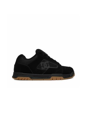 Zapatilla baja negra con cordones negros, suela de goma con dibujo marrón claro y un logo DC gris oscuro en el panel lateral.