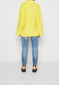 Personne portant un pull jaune, un jean bleu clair et des chaussures à plateforme beiges se tenant contre un fond blanc uni.