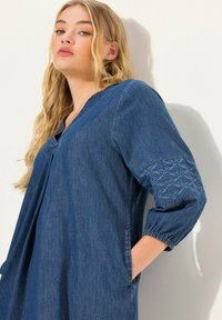 Denimmekko, jossa on V-pääntie, puffihihat ja brodeeratut yksityiskohdat hihoissa. Mukana on kaksi sivutaskua ja rento malli.