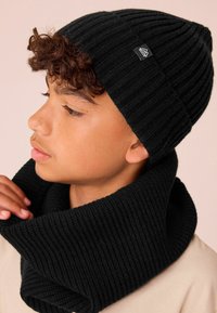 Sort ribbet beanie og halsvarmer lavet af struktureret strikket stof. Beanie'en sidder tæt, mens halsvarmeren hænger løst omkring halsen.