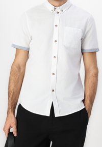 Camicia bianca a maniche corte con polsini azzurri, colletto button-down, bottoni in ottone e una tasca singola sul lato sinistro del petto.