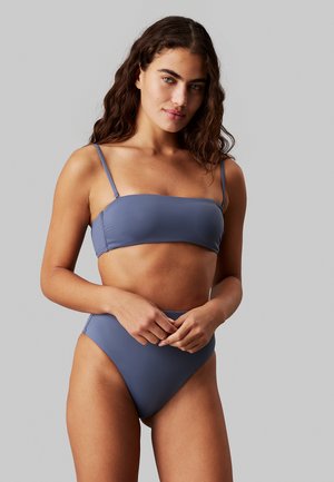 Donna con capelli castani ondulati che indossa un top bikini blu senza spalline e slip bikini a vita alta, in piedi contro uno sfondo grigio.