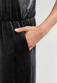 Robe en velours noir avec taille froncée et poches latérales. Texture lisse avec un doux éclat, mettant en valeur une silhouette ajustée.