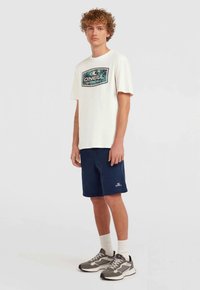 Maglietta bianca con una stampa grafica colorata, abbinata a pantaloni corti blu scuro e sneaker grigi. Caratteristiche includono maniche corte e collo a giro.