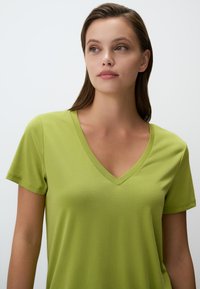 Koszulka z krótkim rękawem w kolorze jaskrawozielonym z dekoltem w kształcie litery V. Wykonana z gładkiego, miękkiego materiału o luźnym kroju. Oferuje minimalne detale i subtelną fakturę.