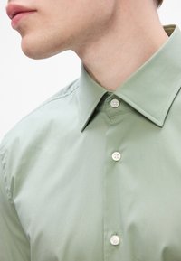 Homem a usar uma camisa verde-clara com botões e colarinho pontudo, mostrado do pescoço até meio do peito contra um fundo branco.