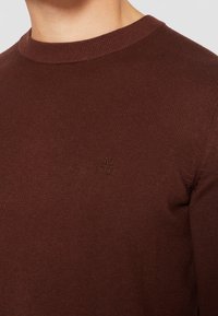 Pull en tricot côtelé marron avec col rond, arborant un petit logo en relief sur la poitrine. Texture douce et coupe ajustée.