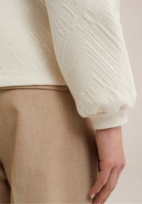 Lähikuva kädestä, joka lepää sivulla, päällä vaalean beige teksturoitu neule, jossa on geometrisia kuvioita, sekä vaaleanbeige housut.