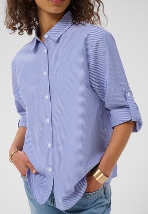 Le modèle porte une chemise boutonnée à rayures bleues et blanches, aux manches retroussées, un jean et des bracelets dorés, et pose les mains dans les poches.
