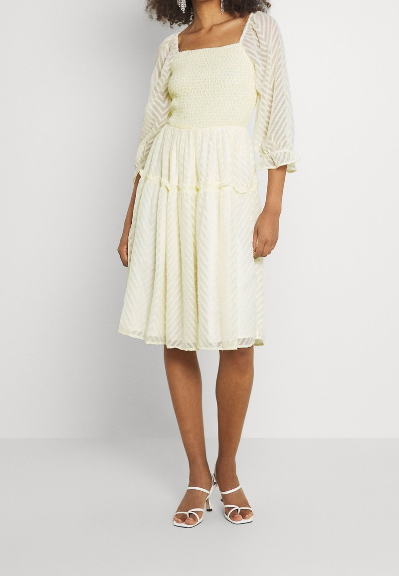 Robe jaune à motifs avec un corsage smocké, des manches trois-quarts transparentes et une jupe à volants. Associée à des talons blancs à lanières.