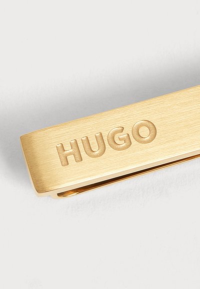 Клипс за вратовръзка от златен метал гравиран с думата HUGO на обикновен бял фон.