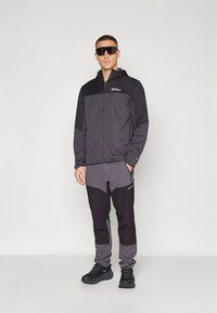 Svart och grå zip-up jacka med hög krage, kombinerad med tajta mörka byxor. Modellen bär mörka solglasögon och svarta skor.