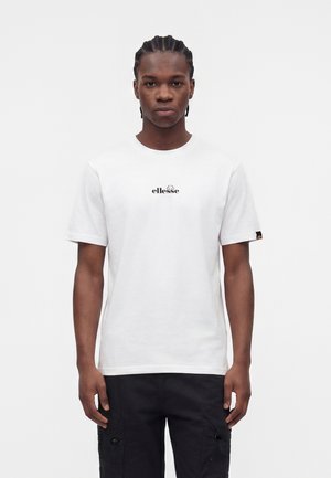 Млад мъж с плитки носи бяла тениска ellesse и черни карго панталони, стоящ срещу обикновен светъл фон.
