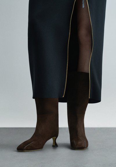 Nő, aki térd fölé érő sötétbarna velúr csizmát visel közepes sarkakkal és fekete szoknyát arany cipzárral.