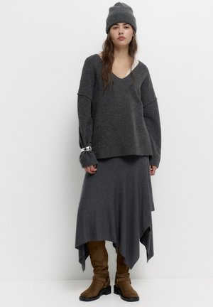 Maglione grigio lavorato a maglia oversize con maniche lunghe, abbinato a una gonna grigia asimmetrica. Stivaletti in camoscio marrone con dettagli in fibbia.