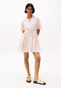 Abito camicia corto rosa chiaro, a maniche corte, con colletto, vita con cordoncino e gonna arricciata. Indossato con sandali scivolati color crema con suola nera.