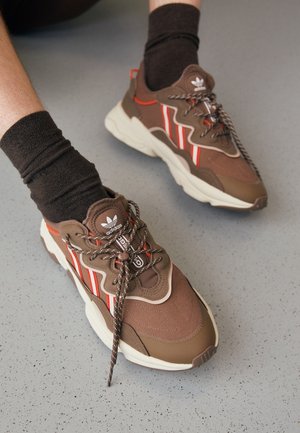 Brązowe sneakersy Adidas z białymi i czerwonymi paskami, noszone z ciemnymi skarpetkami na jasnoszarej, cętkowanej podłodze.