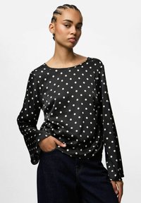 Blusa negra con estampado de lunares blancos, de manga larga y cuello redondo, hecha de un tejido suave. Presenta lunares blancos y un corte holgado, combinada con jeans oscuros.