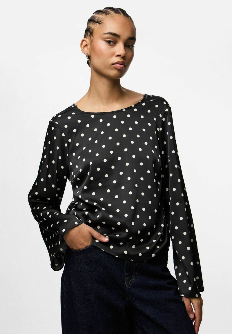 Blusa negra con estampado de lunares blancos, de manga larga y cuello redondo, hecha de un tejido suave. Presenta lunares blancos y un corte holgado, combinada con jeans oscuros.