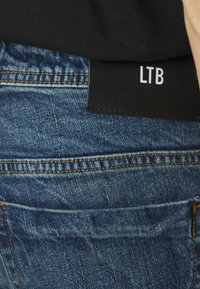 Vaqueros de denim azul con un bolsillo visible y una etiqueta de cuero negro en la cinturilla que dice "LTB". La tela tiene un aspecto texturizado y descolorido.