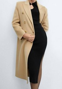 Cappotto doppiopetto in misto lana beige con bottoni dorati sopra un vestito nero in maglia a coste con uno spacco laterale e un dettaglio scollato a chiave.