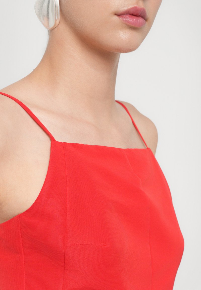 Gros plan sur l'épaule et la mâchoire d'une personne, portant un haut sans manches rouge et de grandes boucles d'oreilles en argent sur un fond clair.