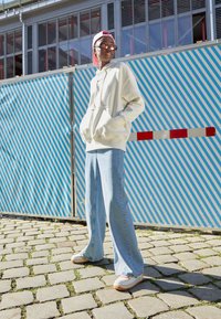 Kremhet genser med snorer, lyseblå vid bukse, og hvite sneakers med fargerike detaljer, satt mot en stripete blå bakgrunn.