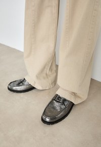 Metallisch grijze loafers met een gemarmerde afwerking, zwarte rubberen zolen en een gestikt ontwerpdetail aan de voorkant, gecombineerd met lichtbeige broeken.