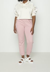 Person klädd i en krämfärgad cropped hoodie, rosa joggerbyxor och vita plattformssandaler med pärldetaljer, stående mot vit bakgrund.