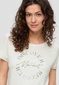 Weißes, kurzärmliges T-Shirt mit rundem Halsausschnitt, das eine metallische, kreisförmige Grafik zeigt, auf der in geschwungenen Buchstaben und Druck steht: "Entdecken Sie den Moment".