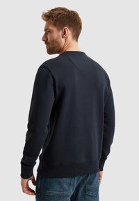 Mornarsko modra bombažna majica s kapuco z okroglim izrezom, rebrastimi manšetami in robom. Na hrbtni strani se nahaja okrogla detajlna maqda.