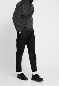 Černé cargo kalhoty se zipovými kapsami, spárované s tmavě šedou poloviční zipovou bundou z měkkého materiálu. Černé boty s texturovanou podrážkou.