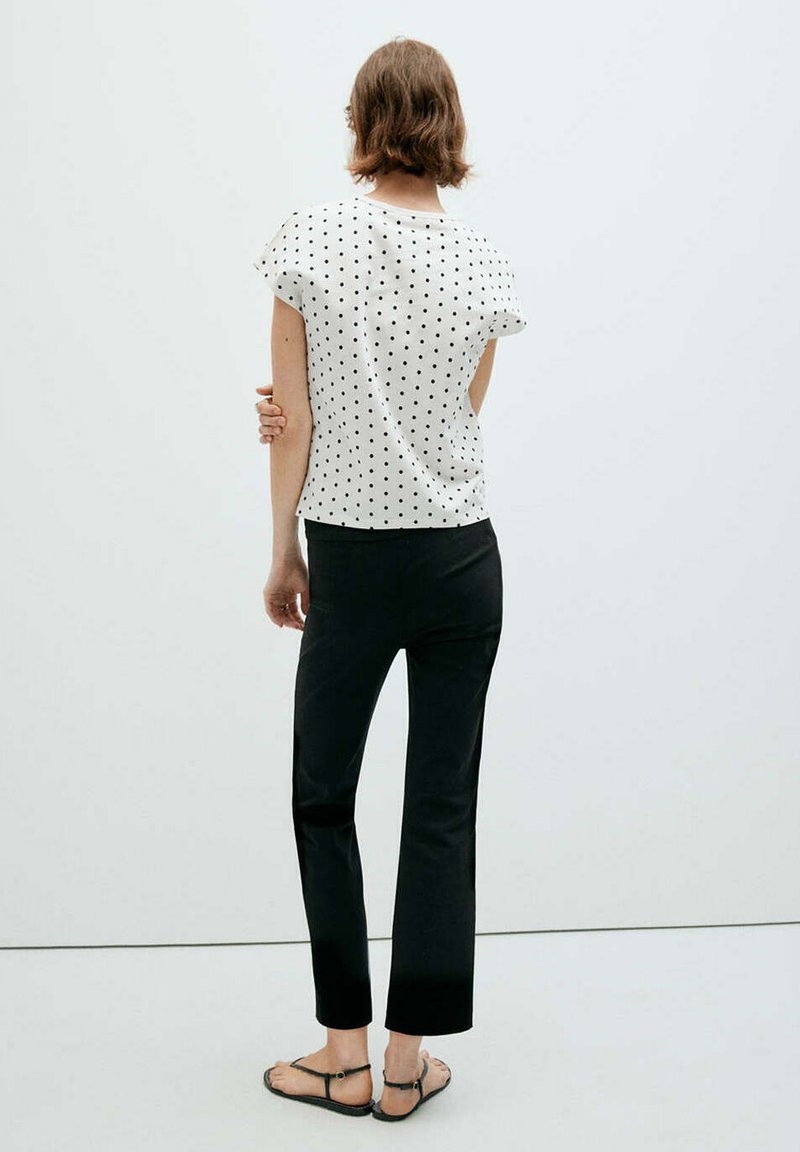 Femme aux cheveux courts et bruns portant une chemise blanche à pois, un pantalon noir et des sandales noires, debout face à un mur blanc, un bras croisé.