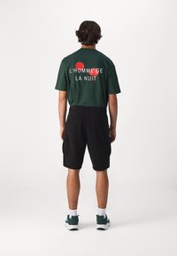 T-shirt verde con testo sul retro "L'HOMME DE LA NUIT" e accenti rossi, abbinato a pantaloni corti neri e scarpe da ginnastica verdi su uno sfondo neutro.