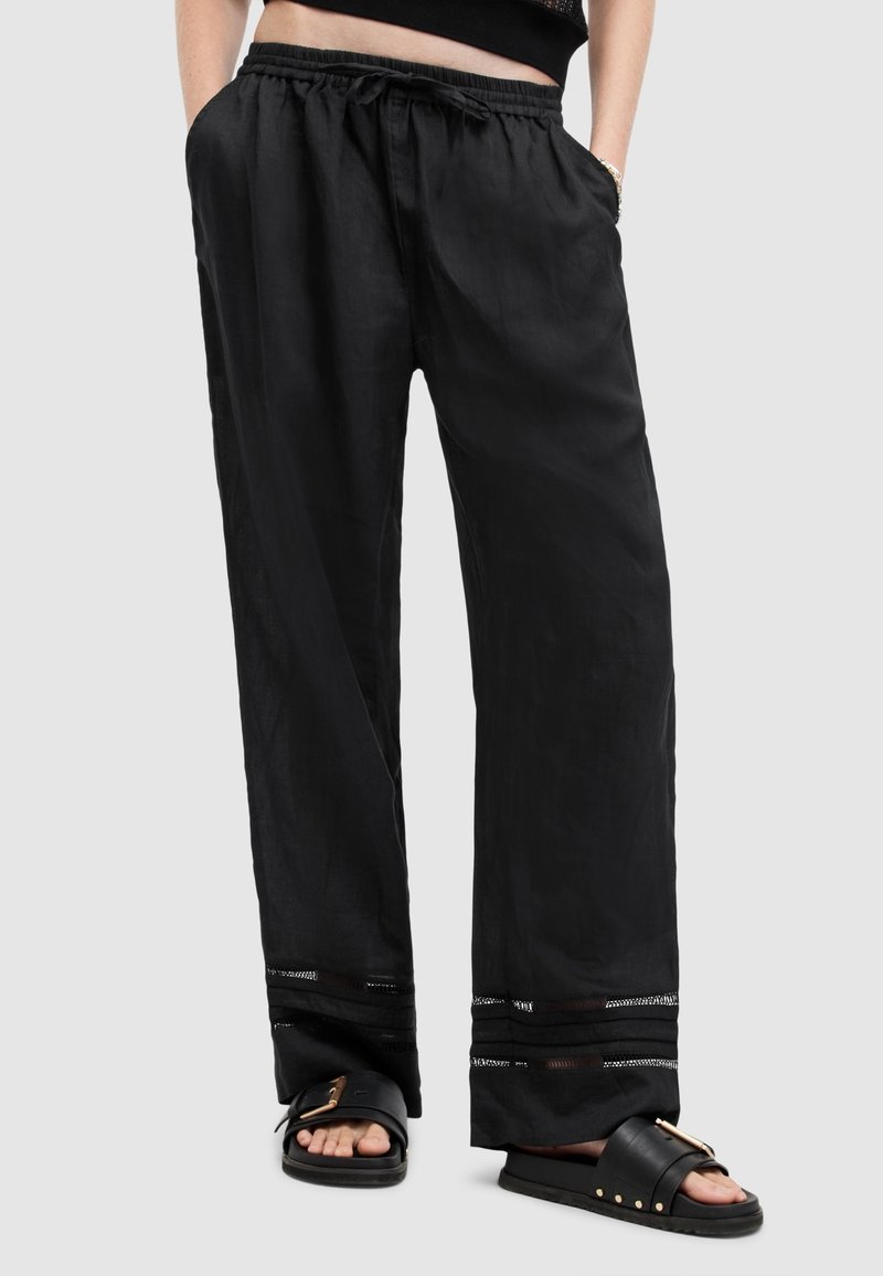 Personne portant un pantalon noir ample à cordon avec des rayures horizontales transparentes près de l'ourlet et des sandales noires à enfiler avec des boucles dorées.