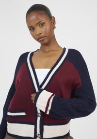 Sweter kardigan w kolorach granatowym, bordowym i kremowym, z dekoltem w kształcie V, w oversize'owym kroju i z zapięciem na guziki. Zawiera kieszenie z przodu z paskami.