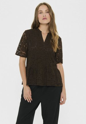 Frau mit langen Haaren, die eine dunkelbraune Spitzenbluse mit kurzen Ärmeln und schwarze Hosen trägt, vor einem schlichten weißen Hintergrund.