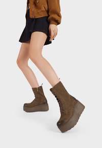 Stivali platform in suede marrone con lacci anteriori, cerniera laterale e suola in gomma spessa, caratterizzati da una punta leggermente arrotondata e una finitura texturizzata.
