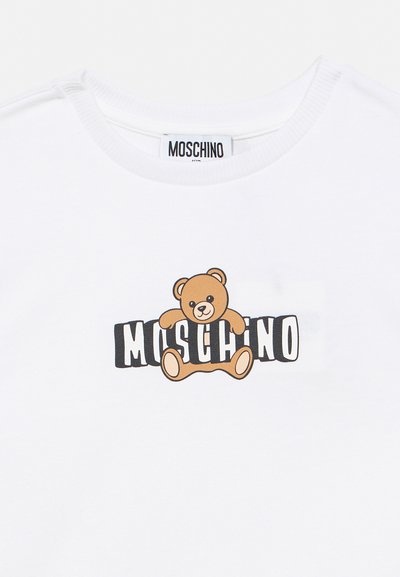 Λευκό μπλουζάκι με στρογγυλή λαιμόκοψη, με γραφικό καφέ αρκουδάκι που κάθεται πάνω από το έντονο μαύρο και λευκό κείμενο "MOSCHINO" στο στήθος.