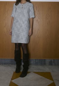 Femme portant une robe à carreaux noir et blanc longueur genoux, des collants noirs et des bottes noires étroites, debout à l'intérieur.