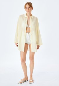 Camicia button-up oversize di un giallo chiaro, con maniche lunghe, indossata sopra un top bikini bianco e pantaloncini bianchi corti. Tessuto morbido e leggero.