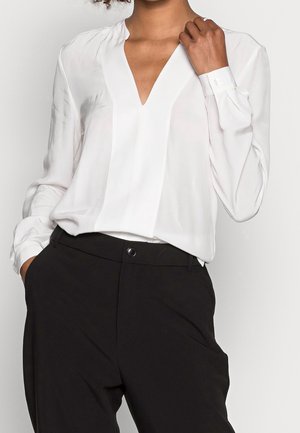 Kvinde iført en hvid langærmet V-hals bluse, der er stoppet ned i højtaljede sorte bukser, med den ene hånd, der rører ved kraven.