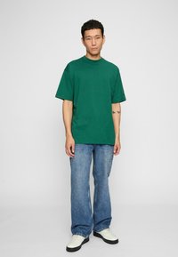 Groen katoenen T-shirt met een relaxed fit en korte mouwen, gecombineerd met losse blauwe denim jeans en witte sneakers. Eenvoudig ontwerp, geen patronen.