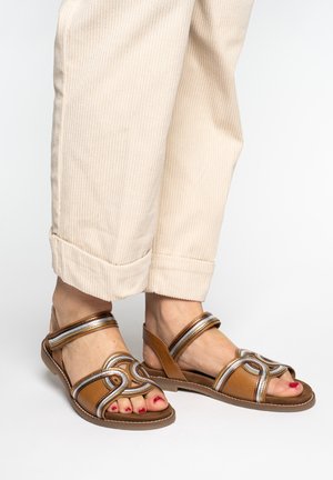 Feet wearing tan leather sandals with silver trim and light beige rolled-up corduroy pants on a white background.